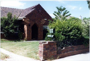 Partial view of varied brown brick house with arched porch with lawn in front of a similar brown brick fence (slightly damaged) in the foreground with decorative metal railing and similar letterbox on the pillar.