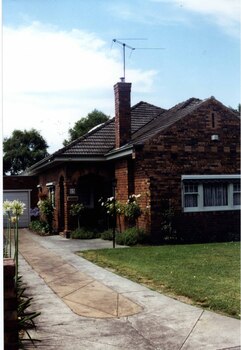 Partial view of mixed brown brick house and garage.  There are a double-arched porch and tall feature chimney with brick patterns plus white windows and white garage door with a concrete drive beside established garden beds and lawn.