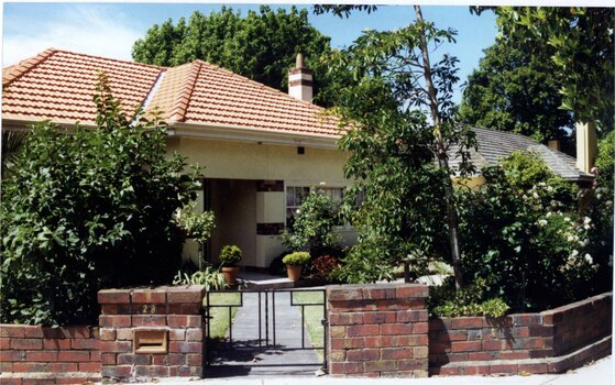 Partial view of front of cream house with brown brick features including on the front porch and chimney, plus low brown brick fence with decorative black metal gate.  Roof and chimney of neighbouring right house just visible through the established garden.  