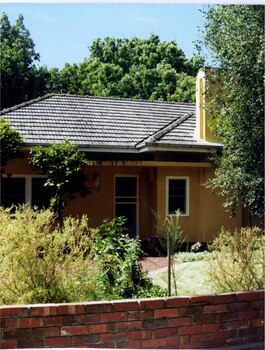 Partial view of tan house and chimney with white framed windows, front door and guttering. Curved low roof jutting over the front door area.  Tall trees behind the house and established plants in the front garden behind the brown brick fence.  