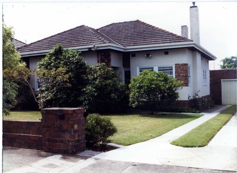 Front and right side of white house with brown brick panel, column and base plus a brown brick garage is partly visible down the drive.  Brown brick fence with established garden behind it.