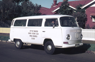 Meals on Wheels delivery van with driver in front of red brick house and white picket fence.
