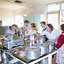 Eight women preparing food around a large table. There are also many large metal containers and a meat slicing machine. 