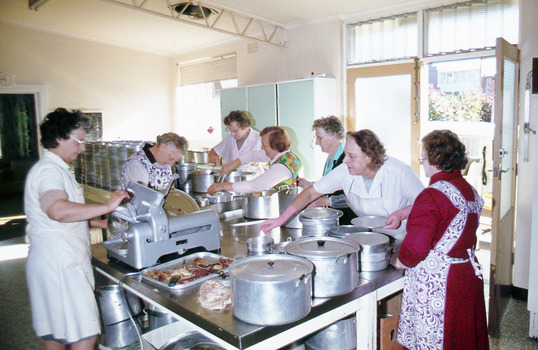 Eight women preparing food around a large table. There are also many large metal containers and a meat slicing machine. 