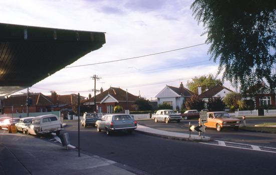 A T intersection with shop awning seen in the foreground and a number of parked cars and cars travelling along the road.