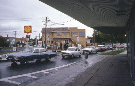 Shell service station on corner of T intersection with cars in the foreground along the road.
