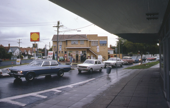 Shell service station on corner of T intersection with cars in the foreground along the road.