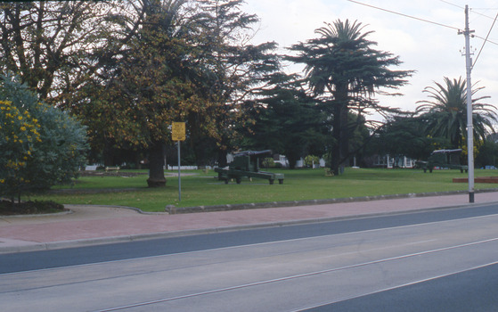 A park with an expanse of lawn, established trees and two canons with a road with tram tracks in the foreground. 