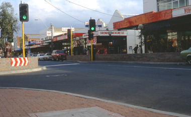 Shopping strip with pedestrian crossing and lights with a curved low brick fence in the foreground.