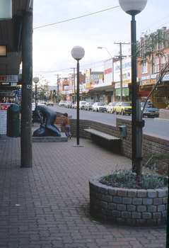 Looking  along shopping strip with a black sculpture and white ball lights on metal poles on paved area in the  foreground.