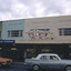 Shopping strip, with Art Deco Façade on upper storey residences. Two cars on the road in front. 