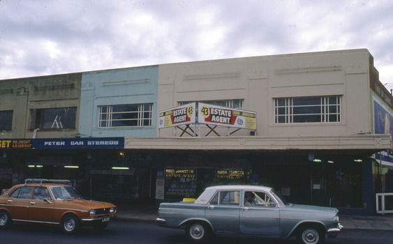Shopping strip, with Art Deco Façade on upper storey residences. Two cars on the road in front. 