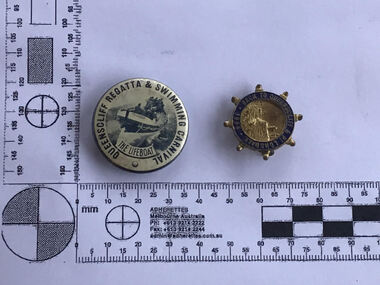 One badge is round on a white base with the inscription 'Queenscliff Regatta & Swimming Carnival' round the edge in dark blue. At the centre of the badge is a photo of the Queenscliff Lifeboat at sea with an inscription reading 'The Lifeboat'. The second badge is shaped like a ships wheel with 8 handles protruding from the centre. The badge is brass coloured with an etched view of a shhip at sea. The blue outer band is inscribed with ' Back to Queenscliff and Pt Lonsdale 1934. 