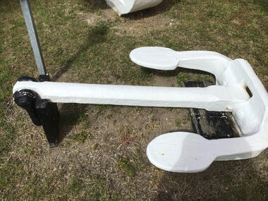 A kedge anchor painted white with two arms with flukes at either end and a flat head with a slot at the centre. The ring at the top is painted black. 