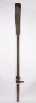 A timber oar with fitted metal rowlocks which has a lower metal strap across the blade attached. The top strap is missing.