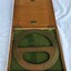 A brass drawing circle protractor in a flat, square wooden box lined with green felt and brass fittings.