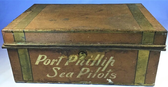 A tin cash box painted brown with two yellow strips painted on either end of it. It contains a lock and key on the front side.