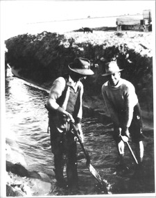 Black & white photo of many two men with shovels digging the channel