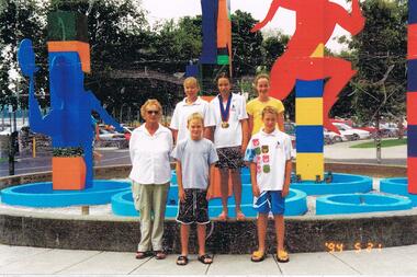 Photograph - VAL CAMPBELL COLLECTION: PHOTOGRAPH OF FIVE CHILDREN AND ONE ADULT IN FRONT OF AN ATHLETICS SCULPTURE, 2003