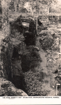 This black-and-white card shows a narrow rocky passage through bushland; on the card is printed “THE ROSE SERIES P. 7557 HYAM PASS, SHOWGROUND RESERVE, NOWRA. COPYRIGHT”.