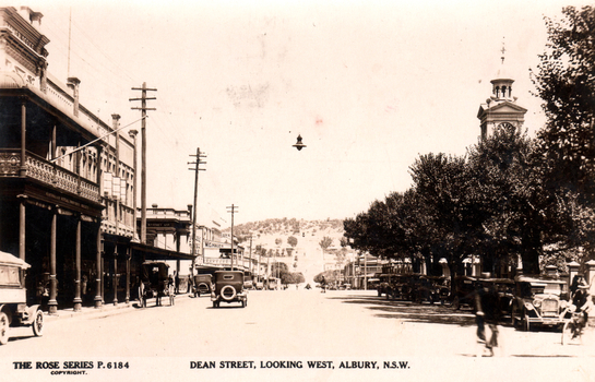 This black-and-white card shows a broad town street lined with shops, motor vehicles and a clock tower; on the card is printed “THE ROSE SERIES P.6184 COPYRIGHT. DEAN STREET, LOOKING WEST, ALBURY, N.S.W.”