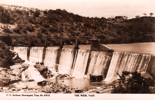 This black-and-white card shows water cascading over a weir in a rural valley; on the card is printed “C. V. Jackson, Newsagent, Yass, No. 6812” and “THE WEIR, YASS”.