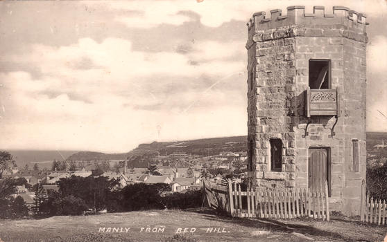 This black-and-white card shows a stone tower overlooking a coastal town; on the card is printed “MANLY FROM RED HILL.”