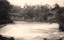 This black-and-white card shows water flowing over a low weir in a rural river valley; there is no visible printed caption.