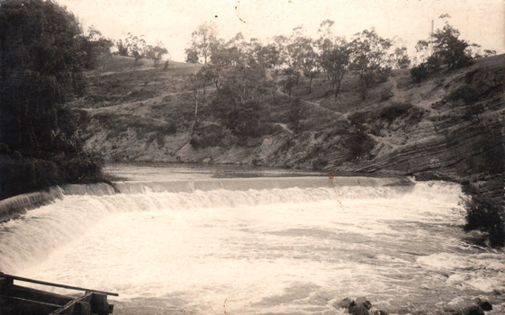 This black-and-white card shows water flowing over a low weir in a rural river valley; there is no visible printed caption.