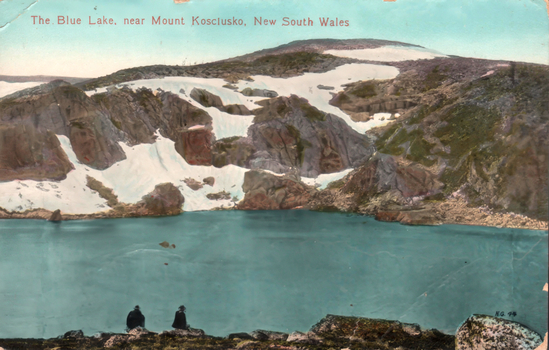 This coloured card shows two people beside a mountain lake surrounded by rocky, snow-covered slopes; on the card is printed “The Blue Lake, near Mount Kosciusko, New South Wales” and “N.G. 44”.