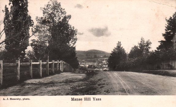 This black-and-white card shows a road descending between trees towards a town; on the card is printed “A. J. Shearsby, photo.” and “Manse Hill Yass”.