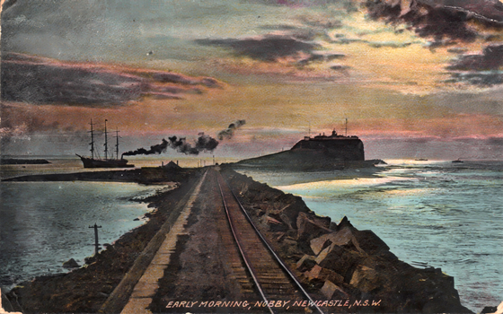 This coloured card shows a coastal railway line leading towards a headland, with a sailing ship offshore; on the card is printed “EARLY MORNING, NOBBY NEWCASTLE, N.S.W.”