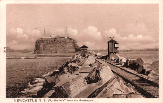 This black-and-white card shows a rocky breakwater with railway tracks leading towards Nobby’s headland: On the card is printed “Ex.195. NEWCASTLE N. S. W. ‘Nobby’s’ from the Breakwater.”