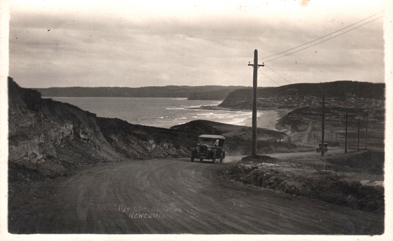 This black-and-white card shows motor cars travelling along a coastal road overlooking the sea; on the card is printed “Memorial Drive Newcastle”.