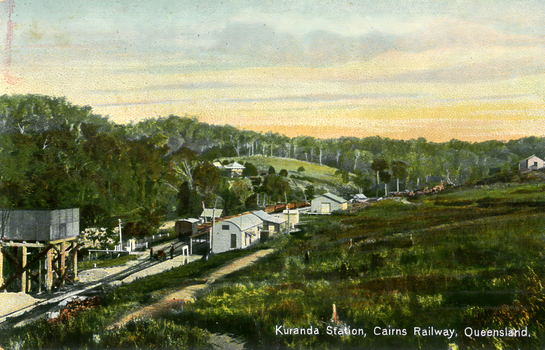 This coloured card shows a rural railway station and tracks surrounded by wooded hills: On the card is printed “Kuranda Station, Cairns Railway, Queensland”.