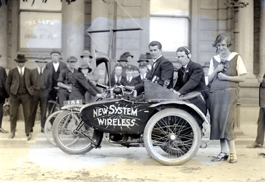 Two men on motor-bike and crowd; watched on by a woman demonstrating mobile radio equipment.