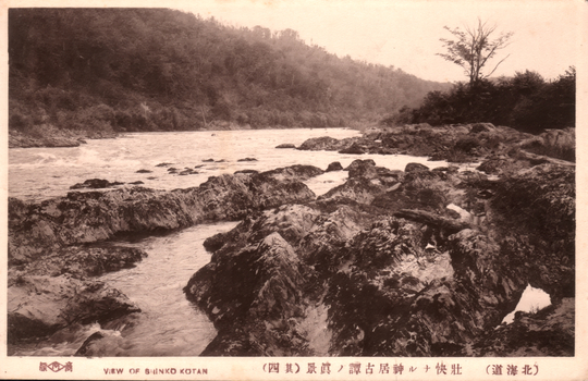 This black-and-white card shows a rocky river flowing through a wooded valley: On the card is printed “商標　VIEW OF SHINKO KOTAN The Japanese is printed mainly in the older right-to-left direction. Read normally:
北海道 — “Hokkaidō.”
壮快ナル神居古譚ノ真景 — “A magnificent true view of Kamui Kotan.”
其四 — “No. 4.”
商標 — “Trademark.”