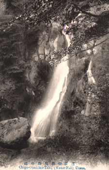 This black-and-white card shows a waterfall descending through a wooded rocky gorge: On the card is printed Chigo-Otosbino-Taki, (Water-Fall) Unzen.” The Japanese is printed in the older right-to-left direction. Read normally, 雲仙嶽稚兒落の瀧 translates as “Chigo-otoshi Waterfall, Mount Unzen.”