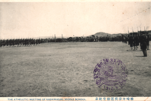 This black-and-white card shows students assembled in formation on a school athletic field. On the card is printed “THE ATHELETIC MEETING OF KASHIWASAKI MIDDLE SCHOOL”. The Japanese is printed from right to left and reads 柏崎中學校運動會紀念, translating as “Commemoration of the Kashiwazaki Middle School athletic meeting.” The circular stamp also shows the date 1912.5; some of its surrounding characters are not clear enough to transcribe confidently.