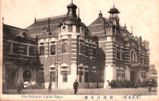 This black-and-white card shows the exterior of a large railway station. On the card is printed “The Shinbashi S at'on Tokyo”. The Japanese is printed from right to left and reads （東京名所）新橋停車場, translating as “Shinbashi Railway Station (a famous place in Tokyo).”