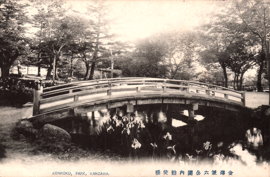 11731.093 This black-and-white card shows an arched wooden bridge over a pond in a landscaped park. On the card is printed “KENROKU, PARK, KANZAWA”. The Japanese is printed from right to left and reads 金澤兼六公園内勅使橋, translating as “Chokushi Bridge in Kenroku Park, Kanazawa.”