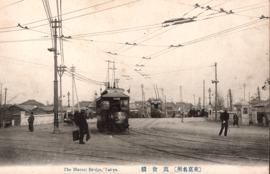 This black-and-white card shows electric trams and pedestrians near a bridge. On the card is printed “The Mansei Bridge, Tokyo”. The Japanese is printed from right to left and reads （東京名所）萬世橋, translating as “Mansei Bridge (a famous place in Tokyo).”