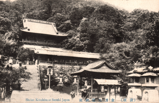 This black-and-white card shows shrine buildings and stone steps on a wooded hillside. On the card is printed “Shrine Kotohira at Sanuki Japan”. The Japanese caption is printed from right to left and reads 金刀比羅神社（讃岐), translating as “Kotohira Shrine, Sanuki.” The sign 本宮 translates as “Main Shrine.”