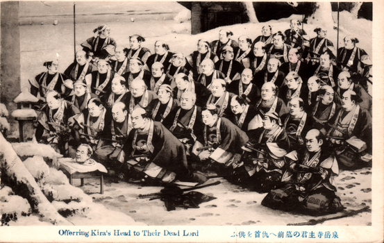 This black-and-white card shows a large group of Japanese men kneeling in a snowy setting around a severed head. On the card is printed “Offerring Kira's Head to Their Dead Lord” 泉岳寺主君の墓前へ仇首を供ふ English translation: “Offering the enemy’s head before their lord’s grave at Sengakuji Temple.”