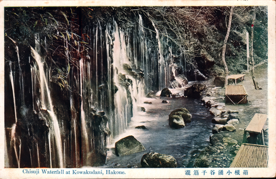 This coloured card shows a waterfall cascading over a rock face into a stream. On the card is printed “Chisuji Waterfall at Kowakudani, Hakone.” The Japanese is printed right-to-left. Read in modern order, it is: 箱根小涌谷千筋瀧 English translation: “Chisuji Waterfall at Kowakudani, Hakone.” 瀧 is the older form of 滝, meaning “waterfall.”