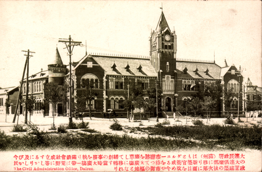 his black-and-white card shows a large civic building with a prominent central clock tower. On the card is printed “The Civil Administration Office, Dairen.” together with a Japanese description in red. The Japanese is printed in the traditional right-to-left direction. The main text begins:
大連民政署（満州）はもとダルニー市廳跡を修繕して創設の事務を執り… and continues by describing the office moving to the Dairen Club, then to a newly constructed government building at the Great Square, and the subsequent development of the surrounding area.
