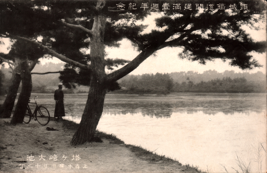 This black-and-white card shows a tree-lined pond or lake, with a man standing beside a bicycle on the shore. The Japanese is printed right-to-left. Read in modern order, it is: 鎭城鐵道開通滿壹週年紀念塔ヶ崎大池上森本驛ヨリ十二丁 English translation: “Commemorating the first anniversary of the opening of the Chinjō Railway.” “Tōgasaki Great Pond.” “Twelve chō from Kamimorimoto Station.”