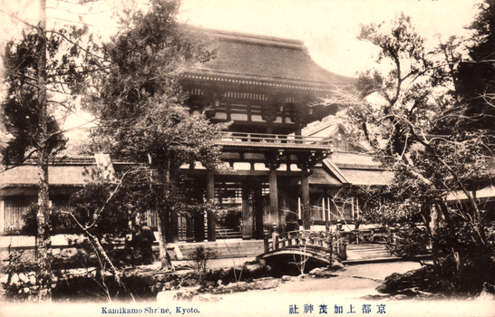 This black-and-white card shows the entrance to a traditional Japanese shrine surrounded by trees and garden paths. On the card is printed “Kamikamo Shr'ne, Kyoto.” The Japanese is printed right-to-left. Read in modern order, it is: 京都上加茂神社 English translation: “Kamigamo Shrine, Kyoto.”