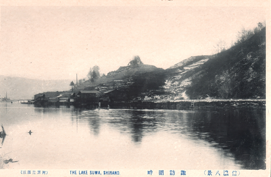 11731.121 This black-and-white card shows Lake Suwa with buildings along the shoreline and wooded hills beyond. On the card is printed “THE LAKE SUWA, SHINANO.” The Japanese translates as:
諏訪湖畔 — “Shore of Lake Suwa” / “Lake Suwa shore”
信濃八景 — “Eight Views of Shinano”
河原書店版 — “Kawahara Bookshop edition”