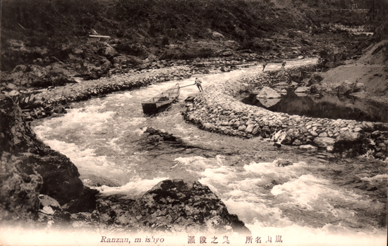 This black-and-white card shows a narrow rocky river channel with rapids, a boat and several people along the bank. On the card is printed “Ranzan, m ishyo”, “嵐山名所” and “奥之段瀬”. The Japanese translates as:
嵐山名所 — “Famous place of Arashiyama”
奥之段瀬 — “Okunodan Rapids” / “the rapids at Okunodan”
